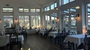 A spacious restaurant with high ceilings and large windows, allowing natural light to fill the room. Tables are neatly arranged, and the floor is wooden.