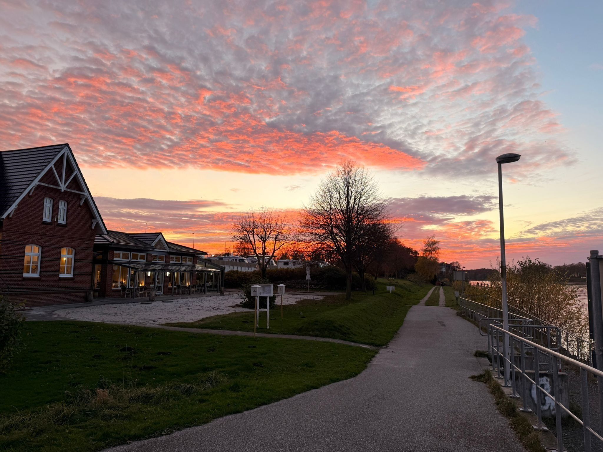 A brick building with a sloped roof and a glass extension, a paved path beside a body of water, and a sunset with pink clouds.