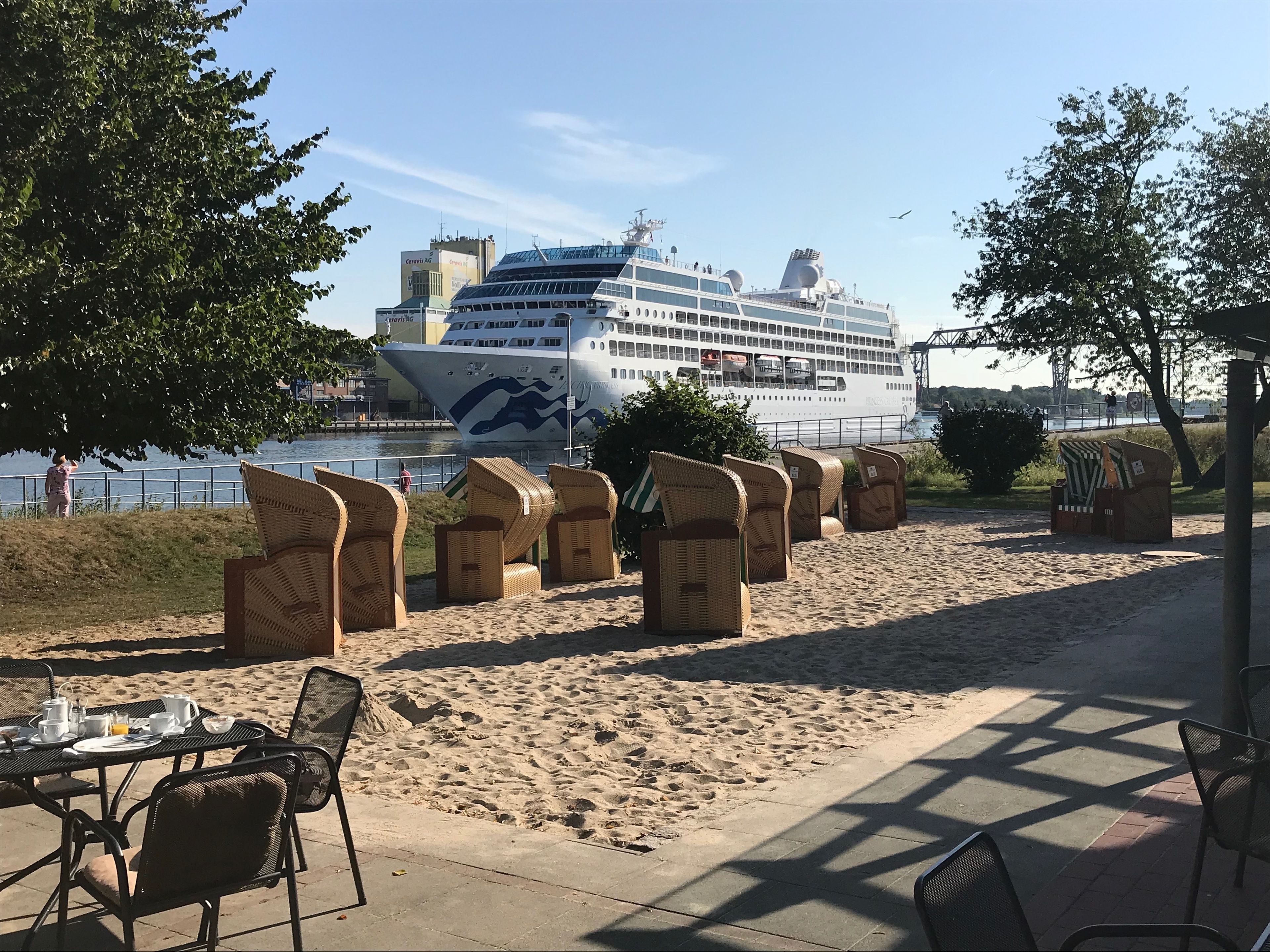 Een eetgedeelte aan het strand met rieten stoelen en tafels, een cruiseschip op de achtergrond en een zandpad.
