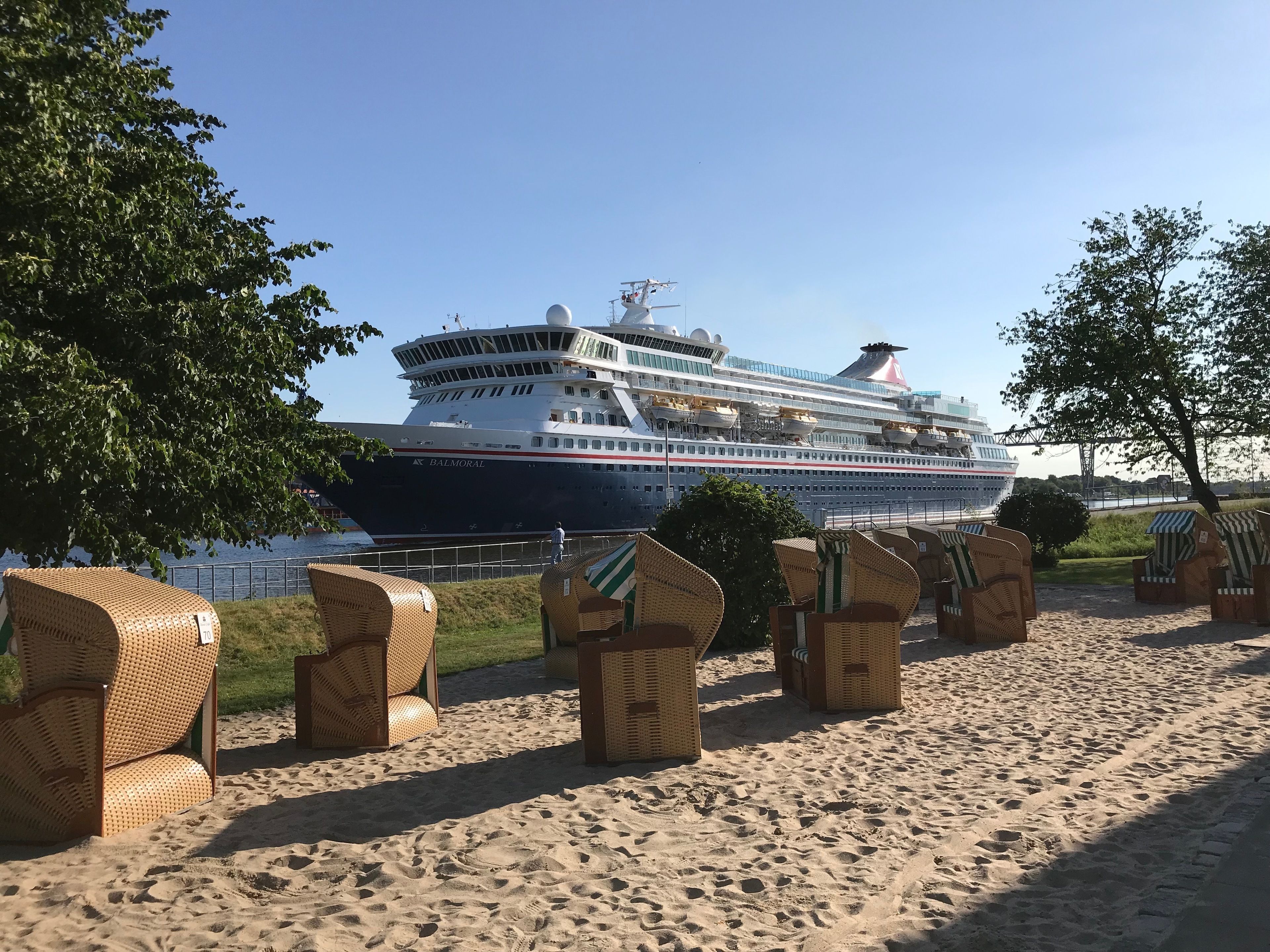 Ett stort kryssningsfartyg låg förtöjt vid en hamn, och i förgrunden fanns det flätade stolar och en sandstrand.