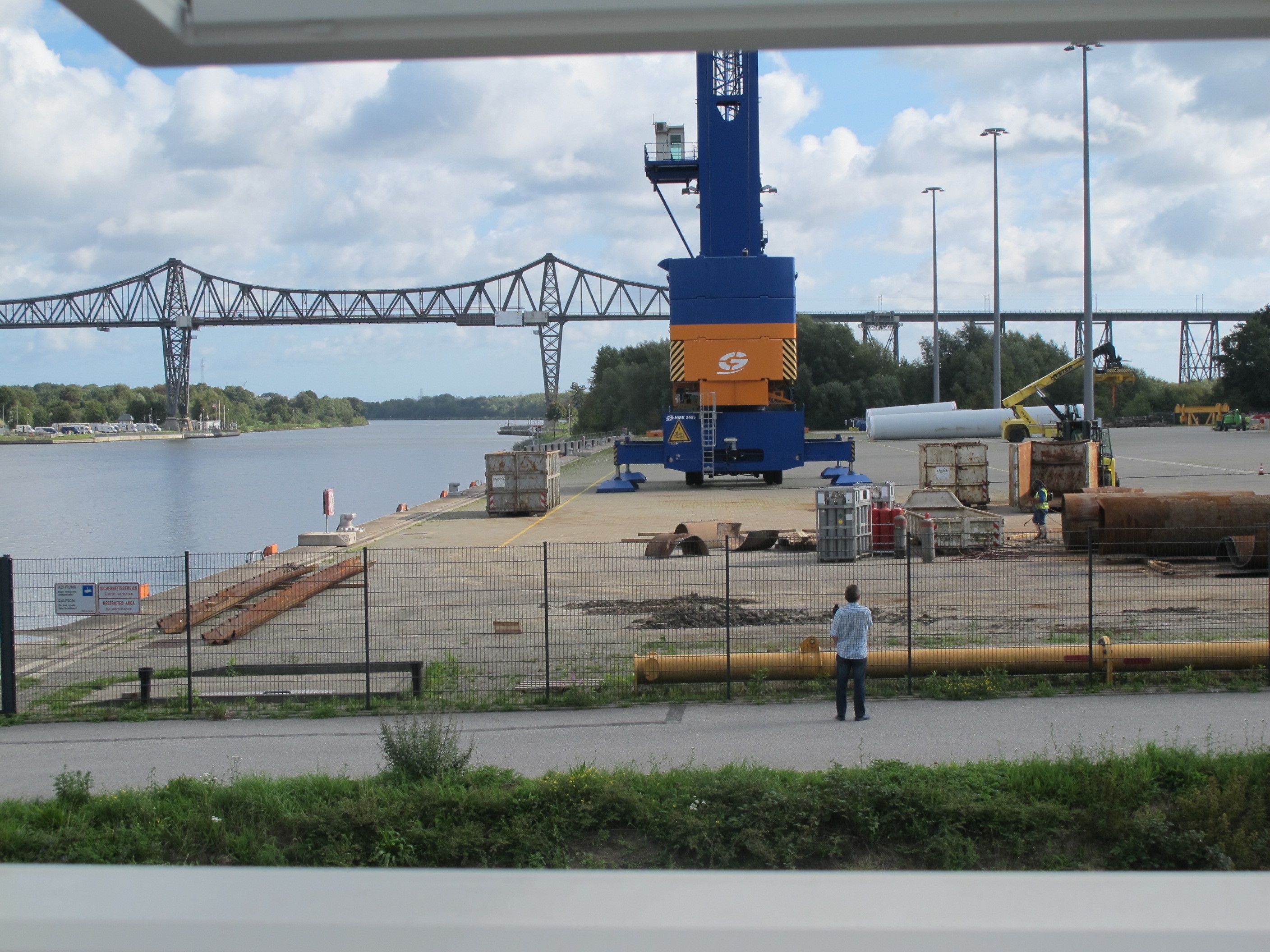 A construction site with a large blue and orange crane, a bridge in the background, and a person standing near a fence.