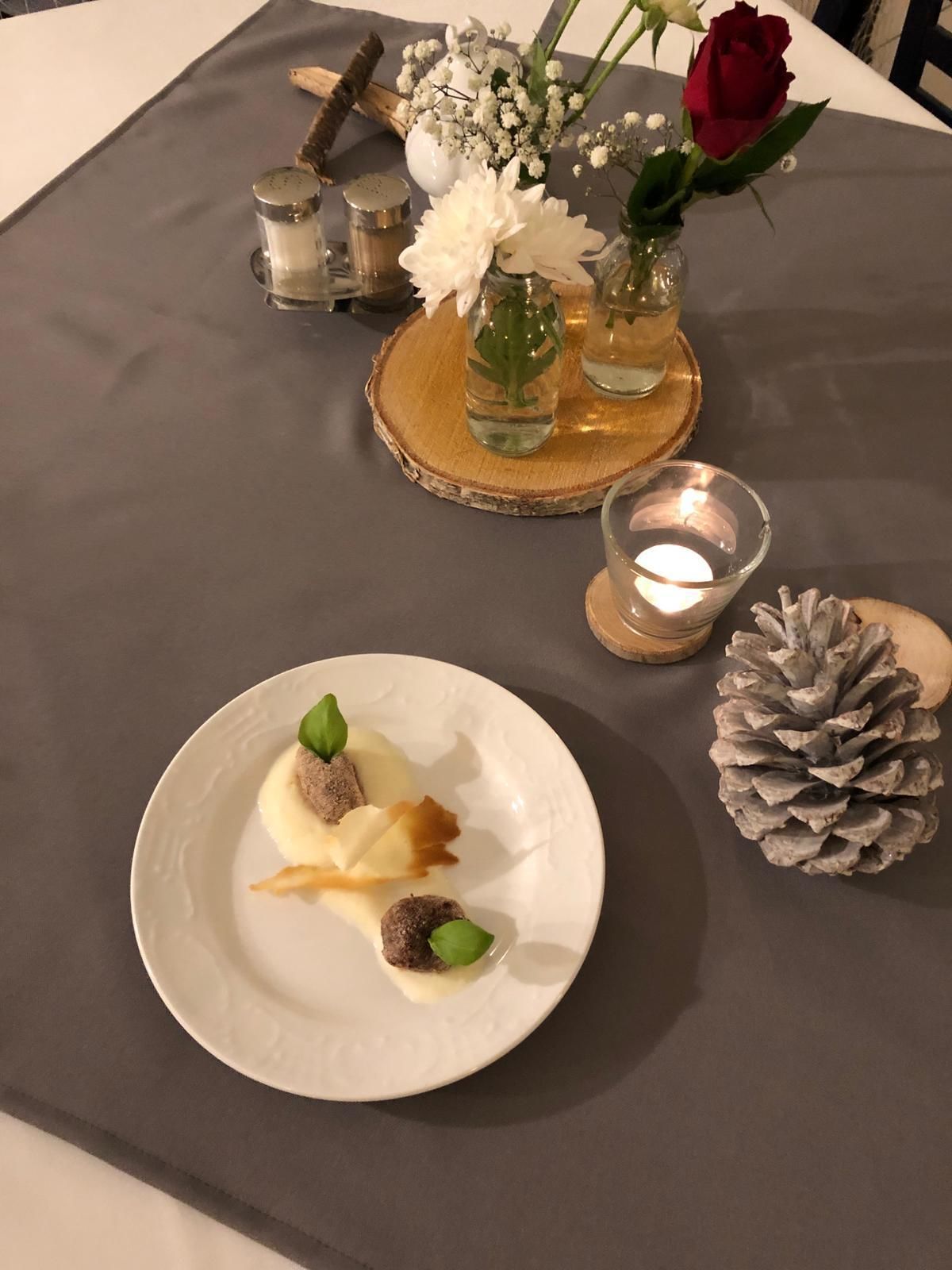 A table set with a white plate of food, a pine cone, a candle, and a vase with flowers and a rose.