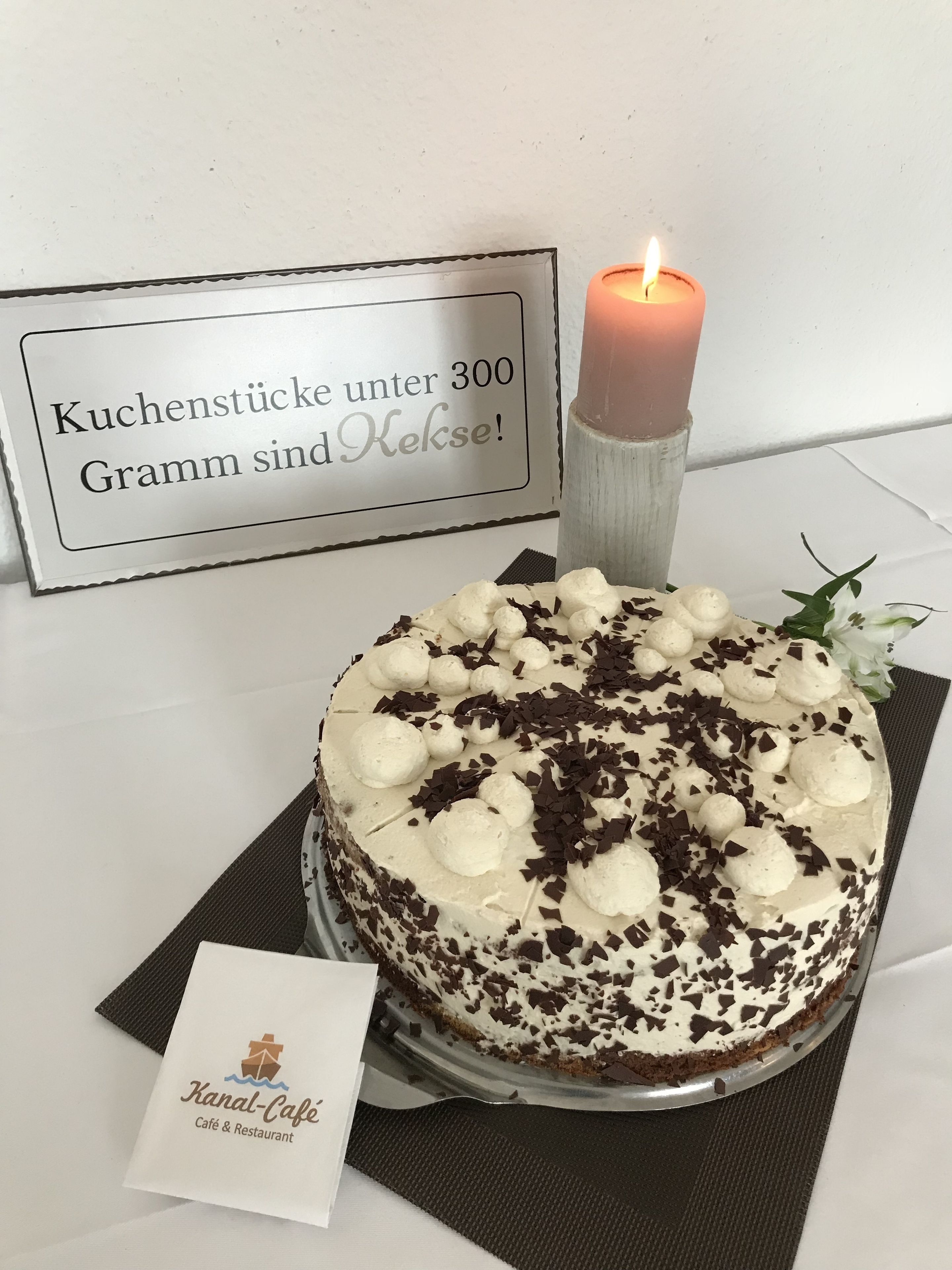 Ein Kuchen mit weißem Zuckerguss und Schokoladenraspeln, eine brennende Kerze und ein Schild mit deutschem Text.