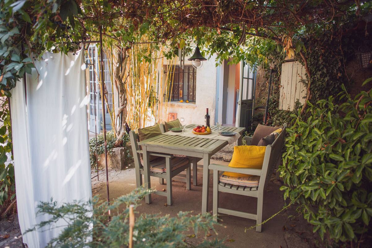 An outdoor patio with a wooden table and chairs, surrounded by greenery and a white curtain.