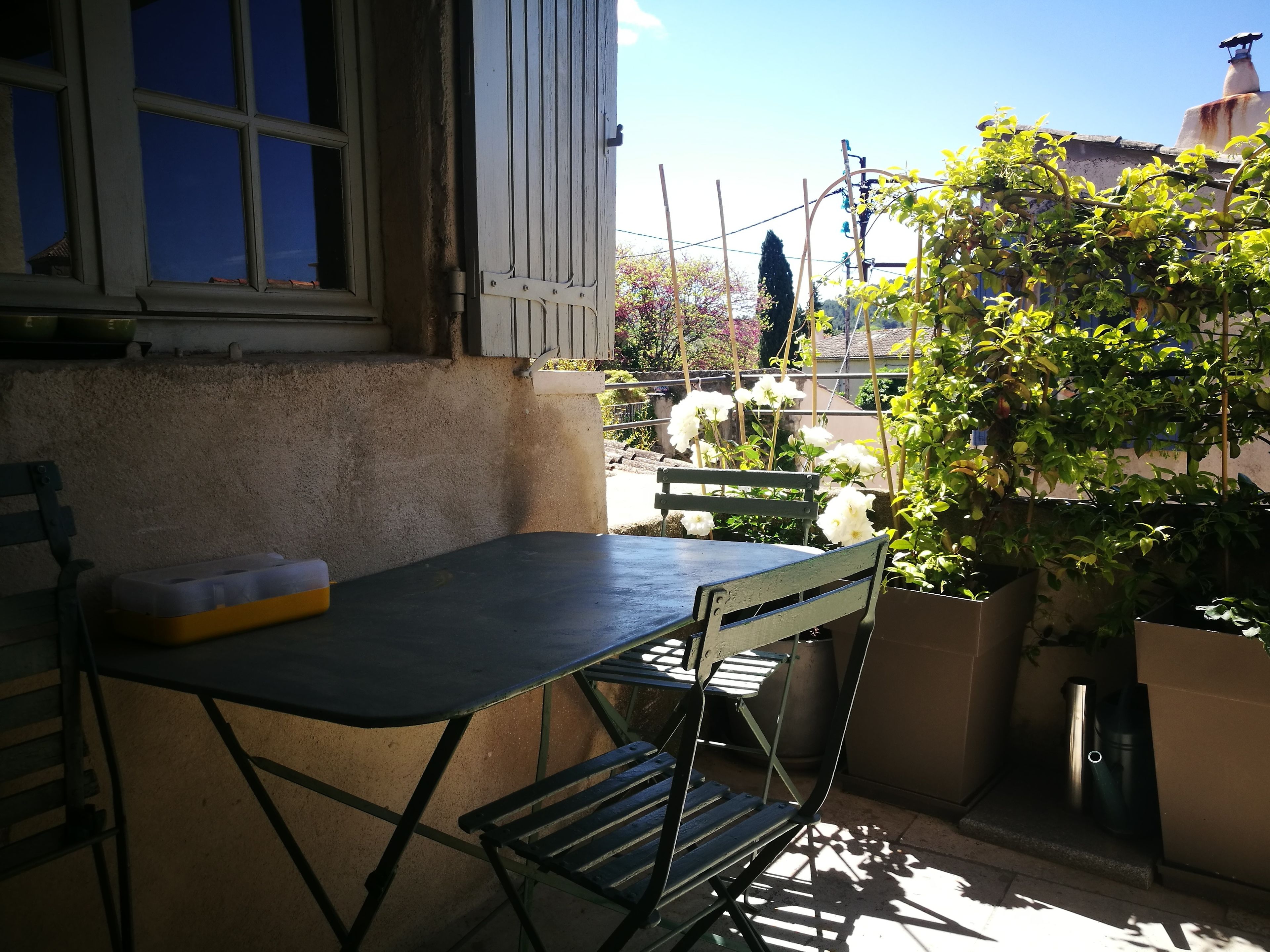 A small outdoor seating area with a table and chairs, a window with shutters, and a potted plant.