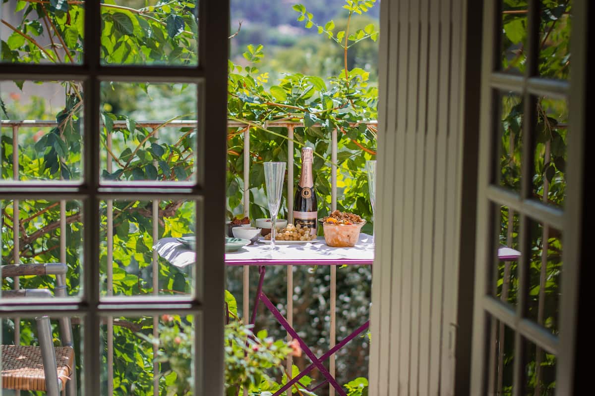 A table with a bottle, bowl, and glass, set against a backdrop of greenery and a window with a grid pattern.