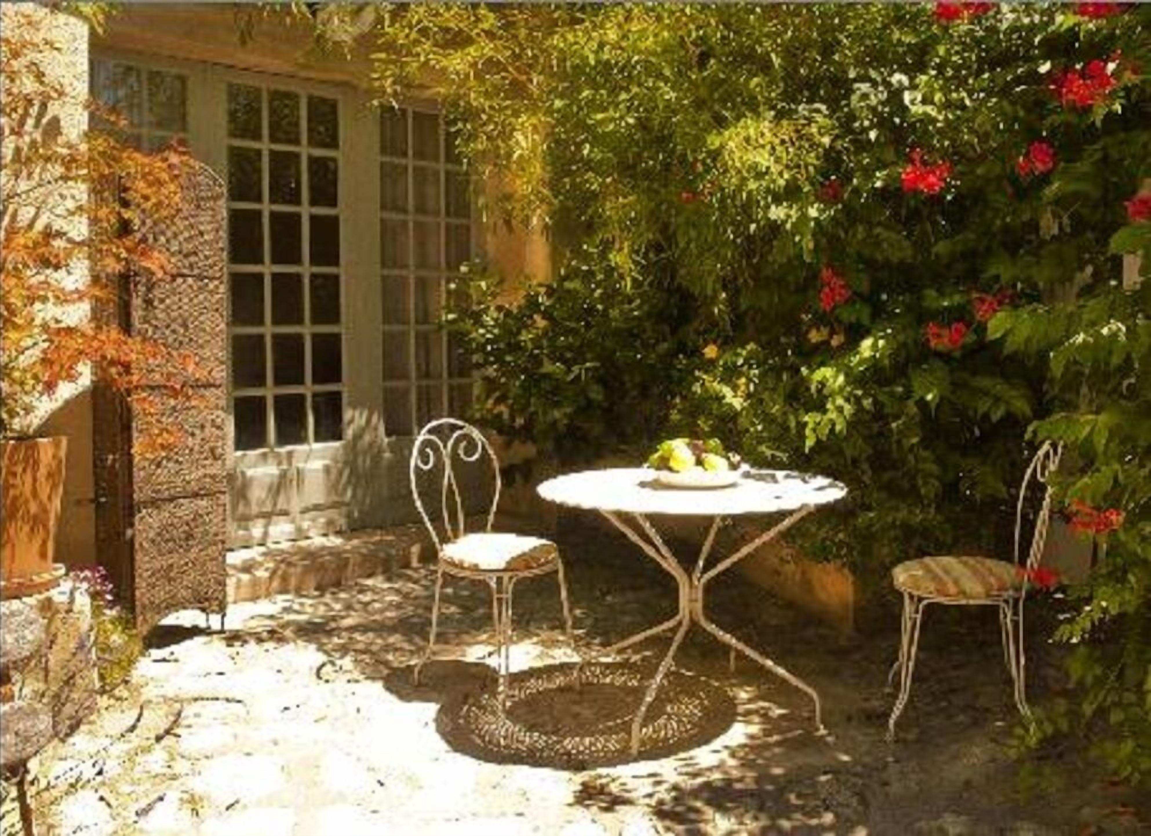 A garden patio with a round table and two chairs, surrounded by lush greenery and a stone wall.