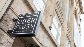 Skilt for 'UBER FLUSS APARTMENTS' med en bygningsfacade i sten i baggrunden.