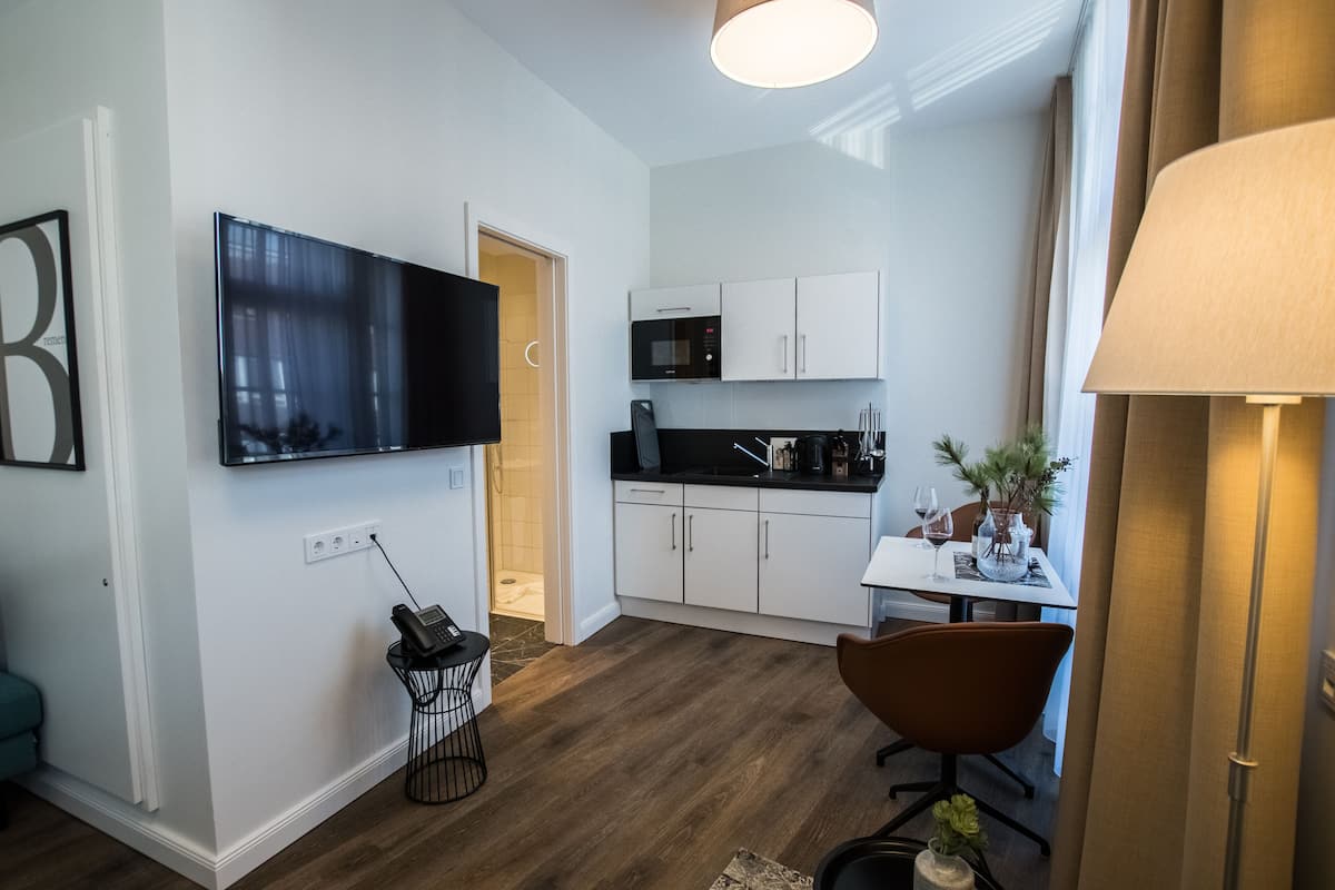 Una cocina moderna con armarios blancos, encimera negra y microondas. Hay un comedor con mesa y sillas, y un televisor montado en la pared.