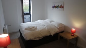 A bedroom with a bed, a bedside table with a lamp, a radiator, and a window with a view of the outside. - Jaylets Premier Apartments, Leicester. (Leicester)