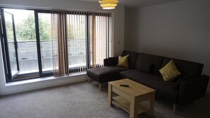 A living room with a sofa, ottoman, and a small table. There is a window with curtains and a view of the outside. - Jaylets Premier Apartments, Leicester. (Leicester)