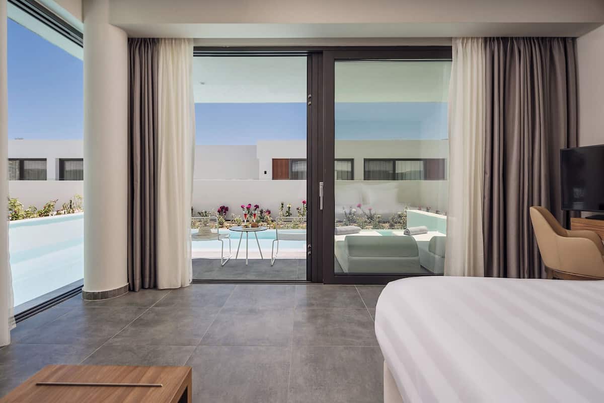 A hotel room with a bed, a TV, a small table, and a view of a pool and outdoor seating area.