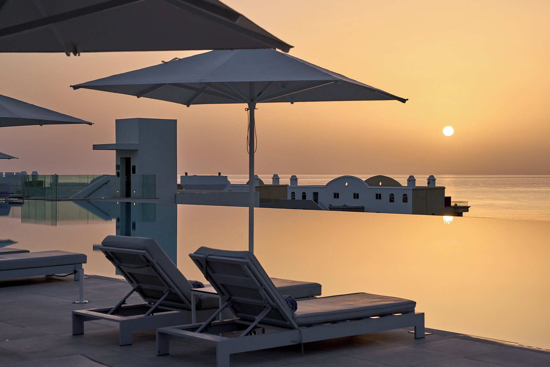 A serene poolside area with lounge chairs and umbrellas, overlooking a calm sea at sunset.