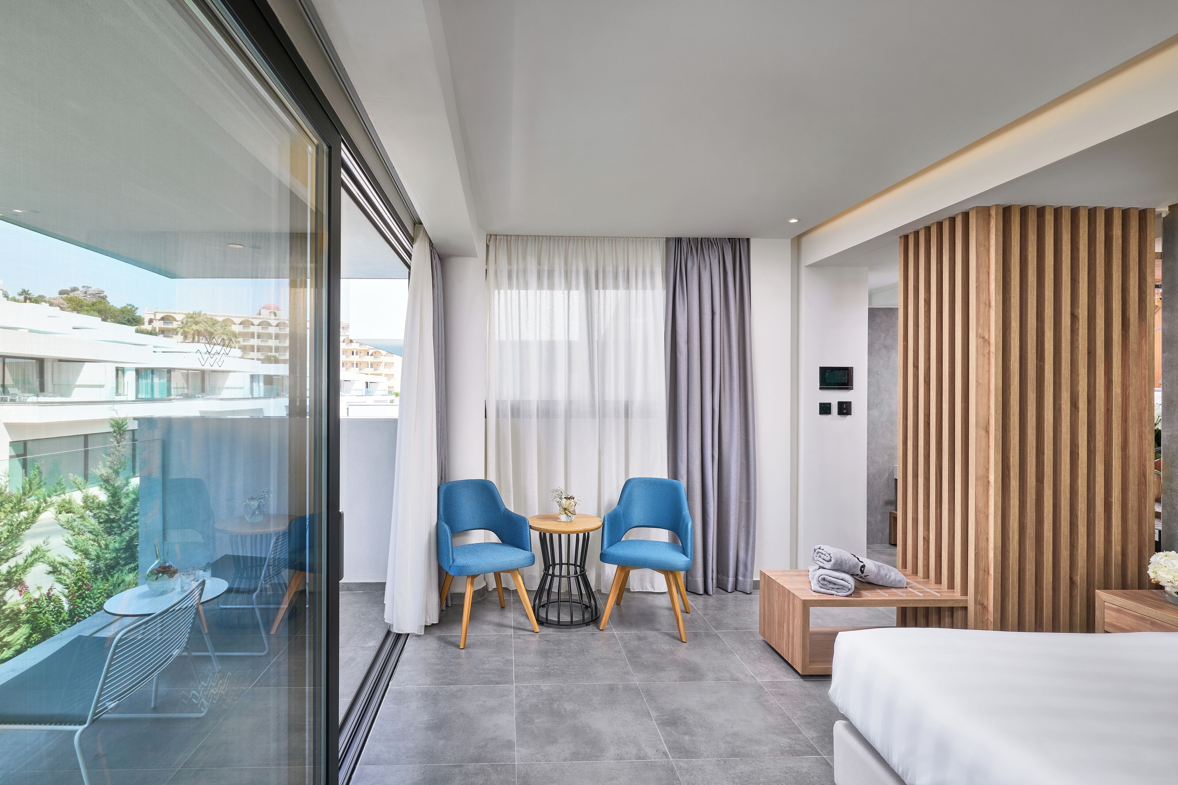 Ein modernes Hotelzimmer mit Balkon, zwei blauen Stühlen, einem kleinen runden Tisch, einem Bett und einer Wandpaneele aus Holz.