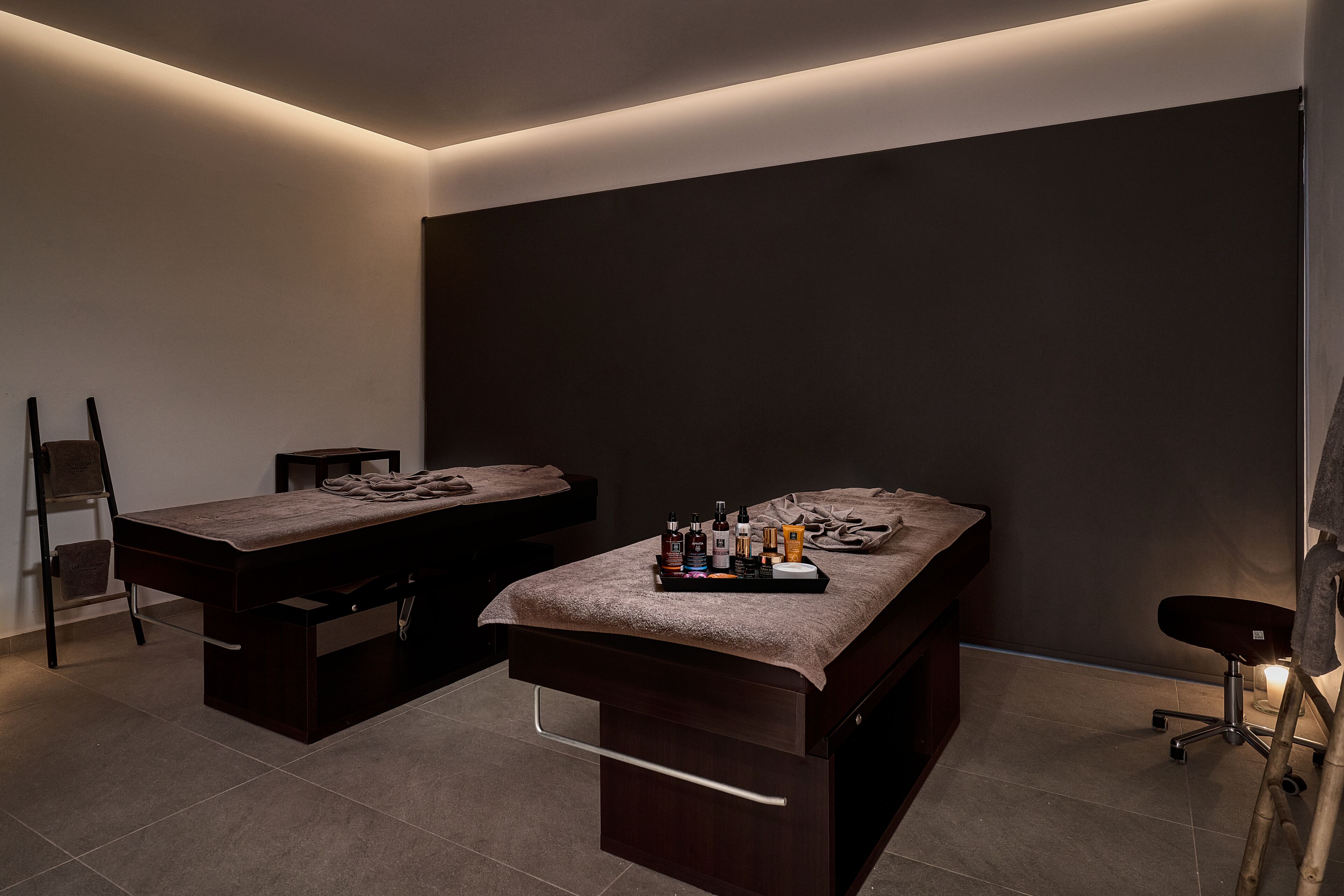 A modern spa room with two massage tables, a small table with bottles, and a chair.