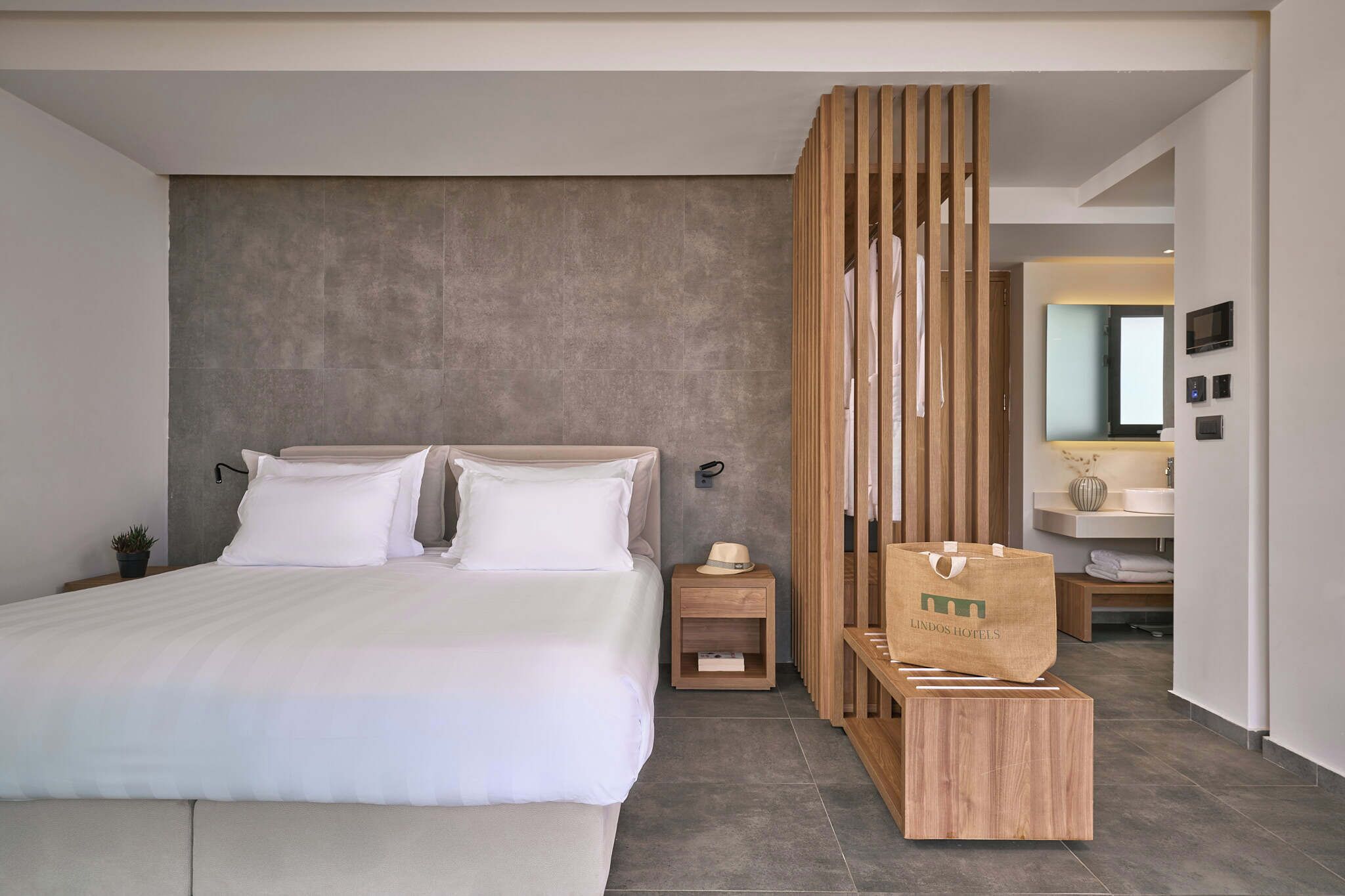 Una habitación de hotel moderna con una cama grande, un separador de madera, una mesita de noche pequeña y un baño visible al fondo.