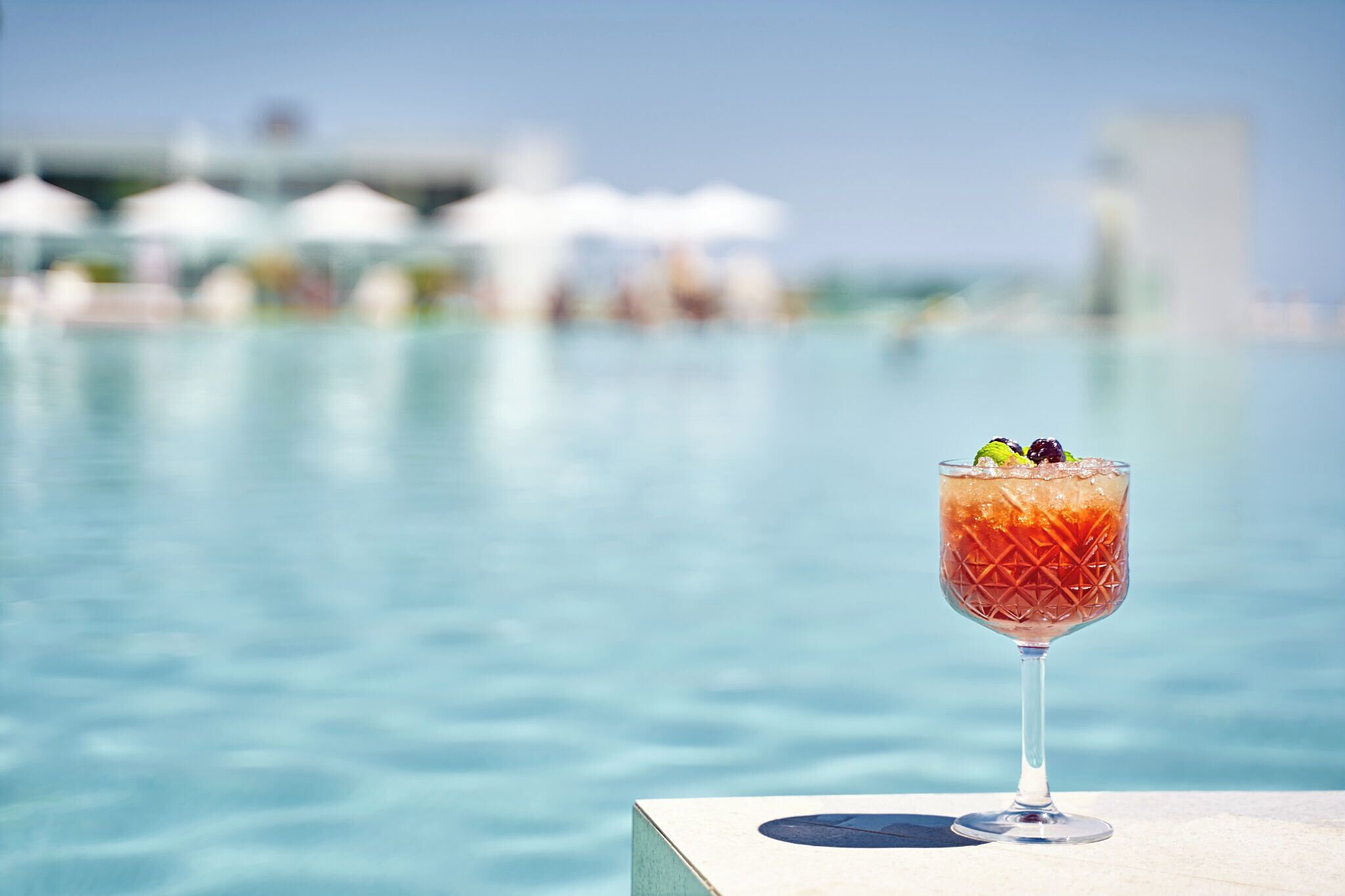A cocktail glass with a garnish on a poolside table, with a pool and umbrellas in the background.