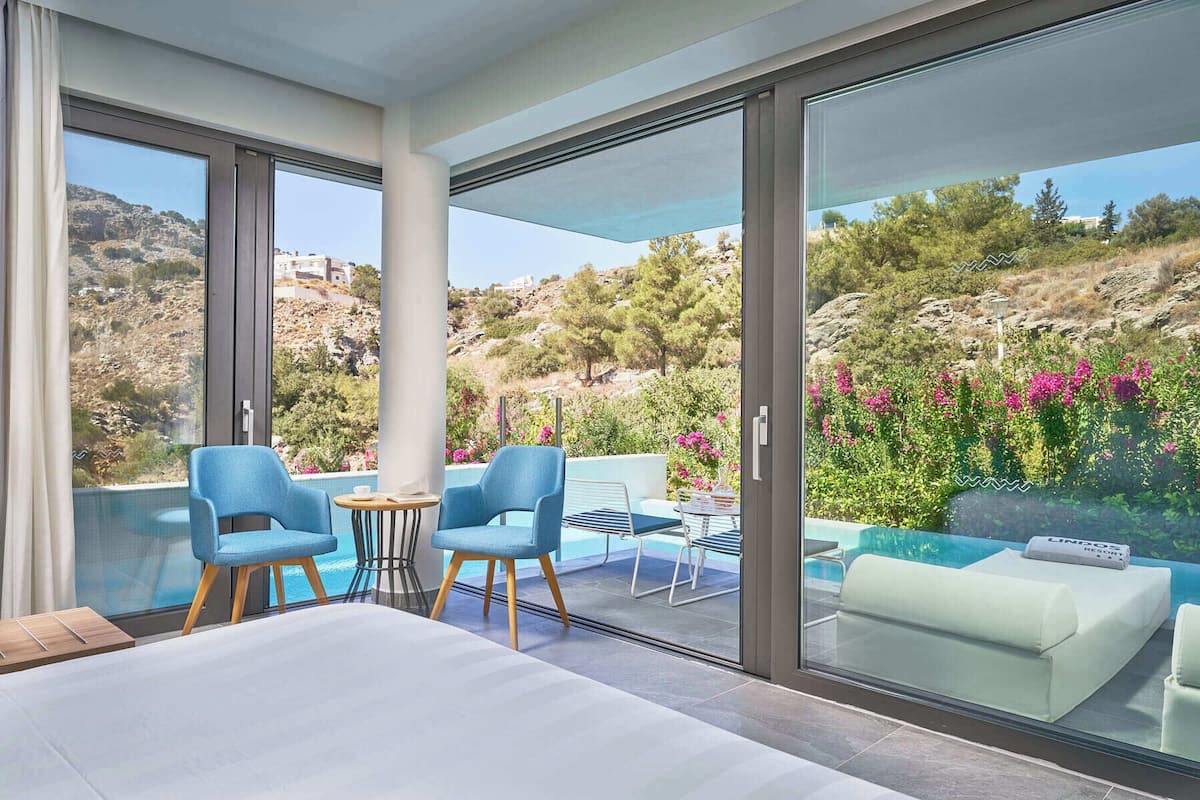 A modern room with a large bed, two blue chairs, a small table, and a sofa. It features large glass doors leading to a balcony with a pool and a view of a hilly landscape.