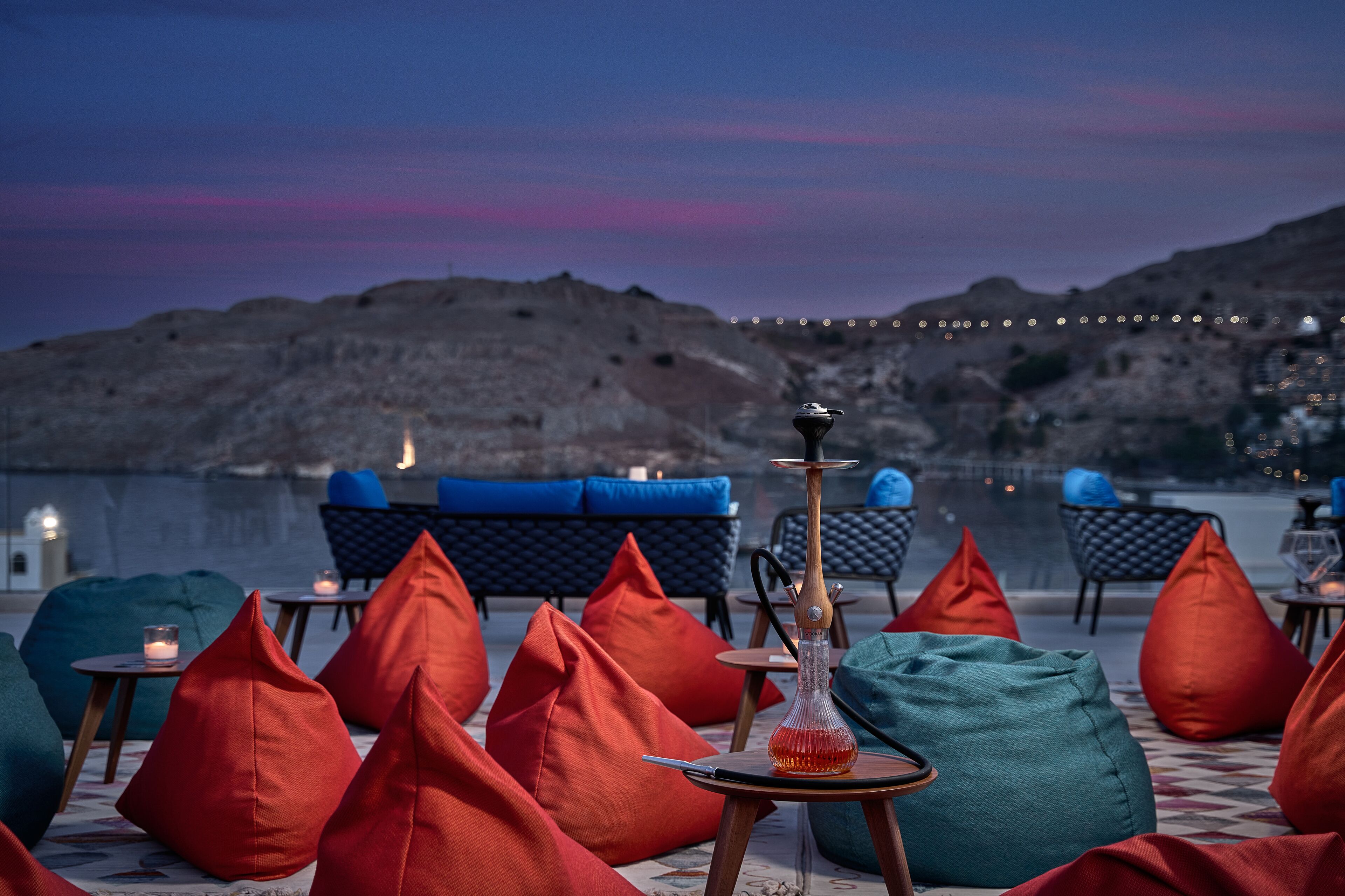 A terrace with outdoor seating, a hookah, and a view of the sea at dusk.