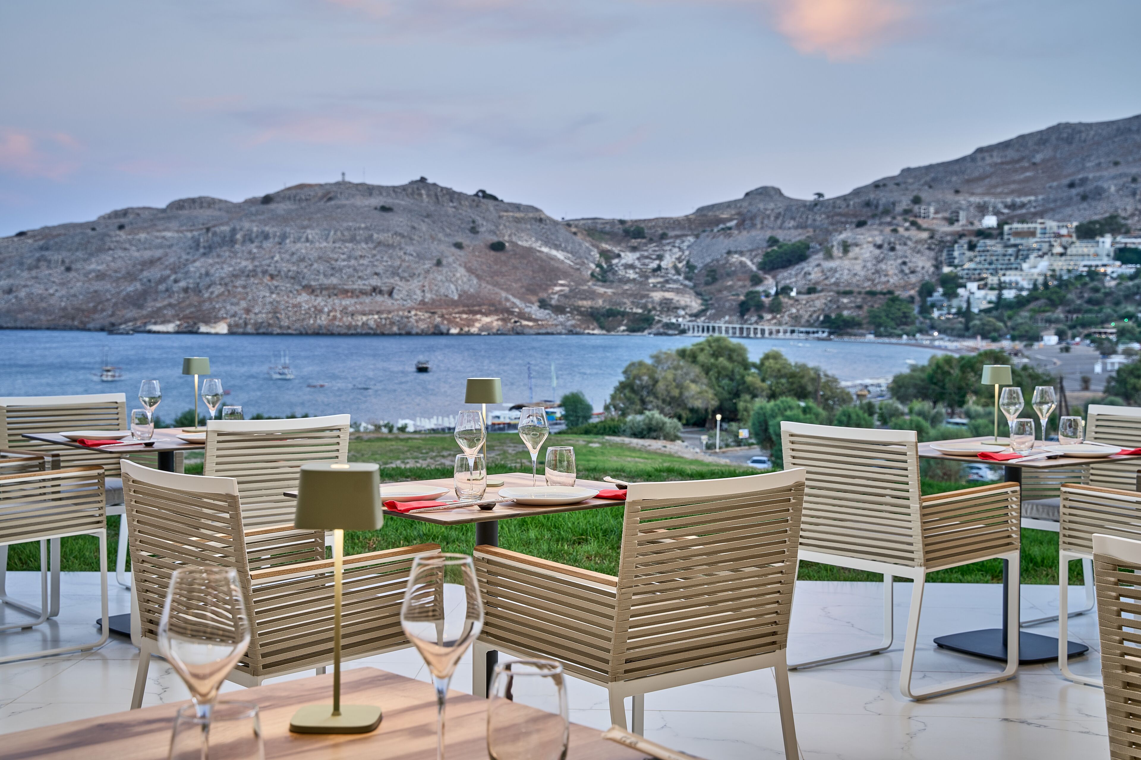 An outdoor dining area with tables and chairs, overlooking a body of water and a hillside town.