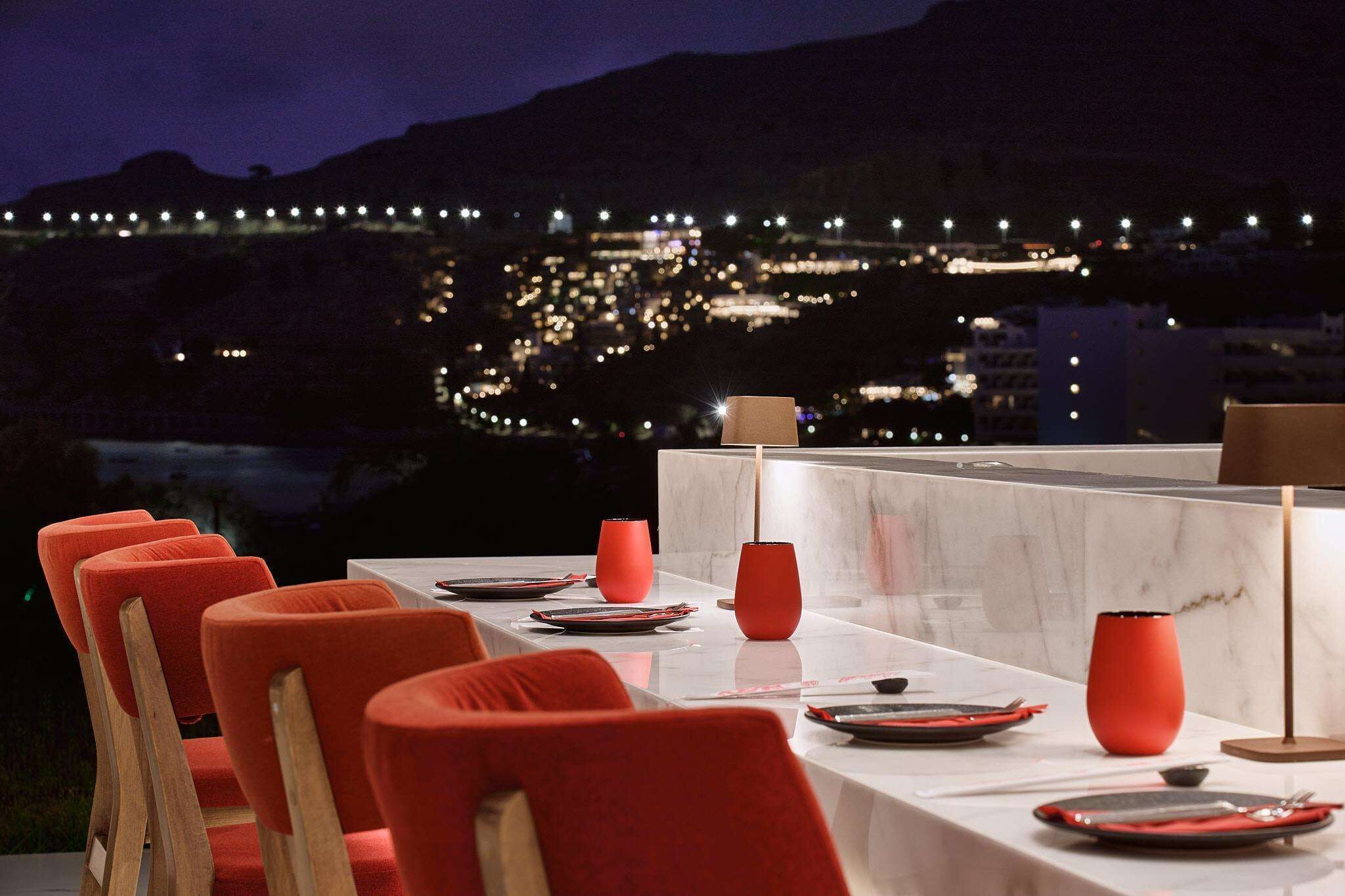 Un coin repas avec une table dressée pour six personnes, des chaises rouges et une vue sur le paysage urbain illuminé la nuit.