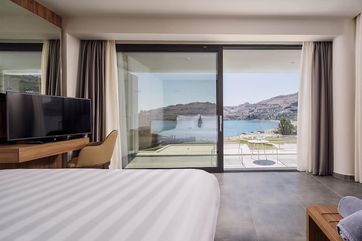 A modern hotel room with a large bed, a flat-screen TV, a glass door leading to a balcony with a pool and scenic views.