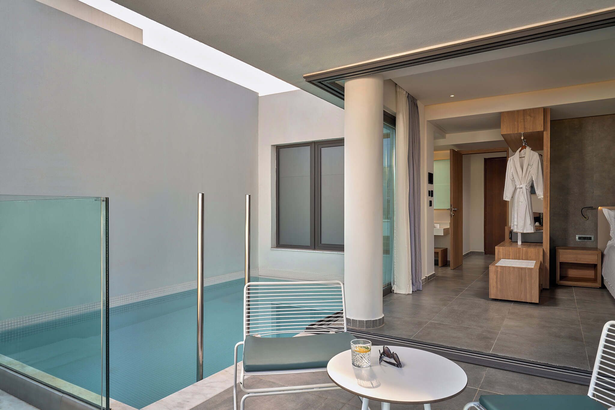 A modern hotel room with a balcony overlooking a swimming pool, a small table with a glass and sunglasses, and a robe hanging on a hook.