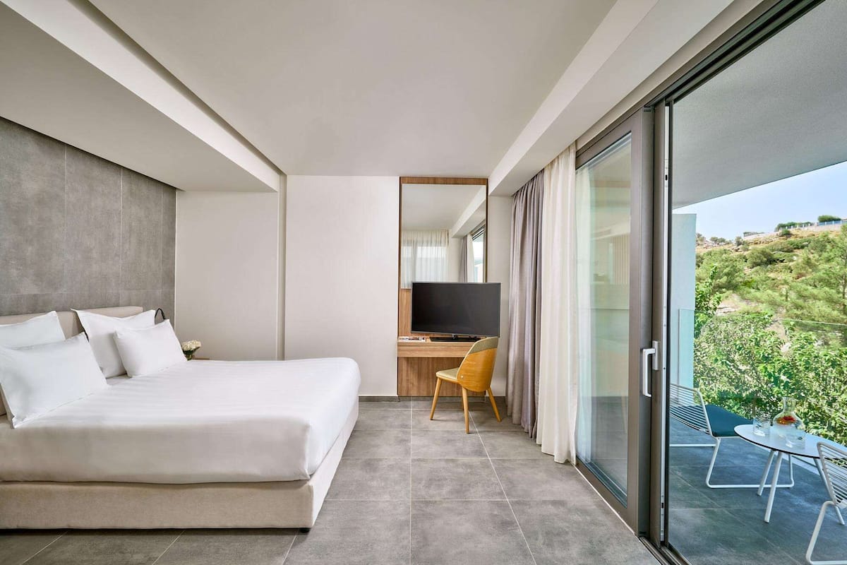 A modern hotel room with a large bed, a desk, and a balcony with a view of greenery.