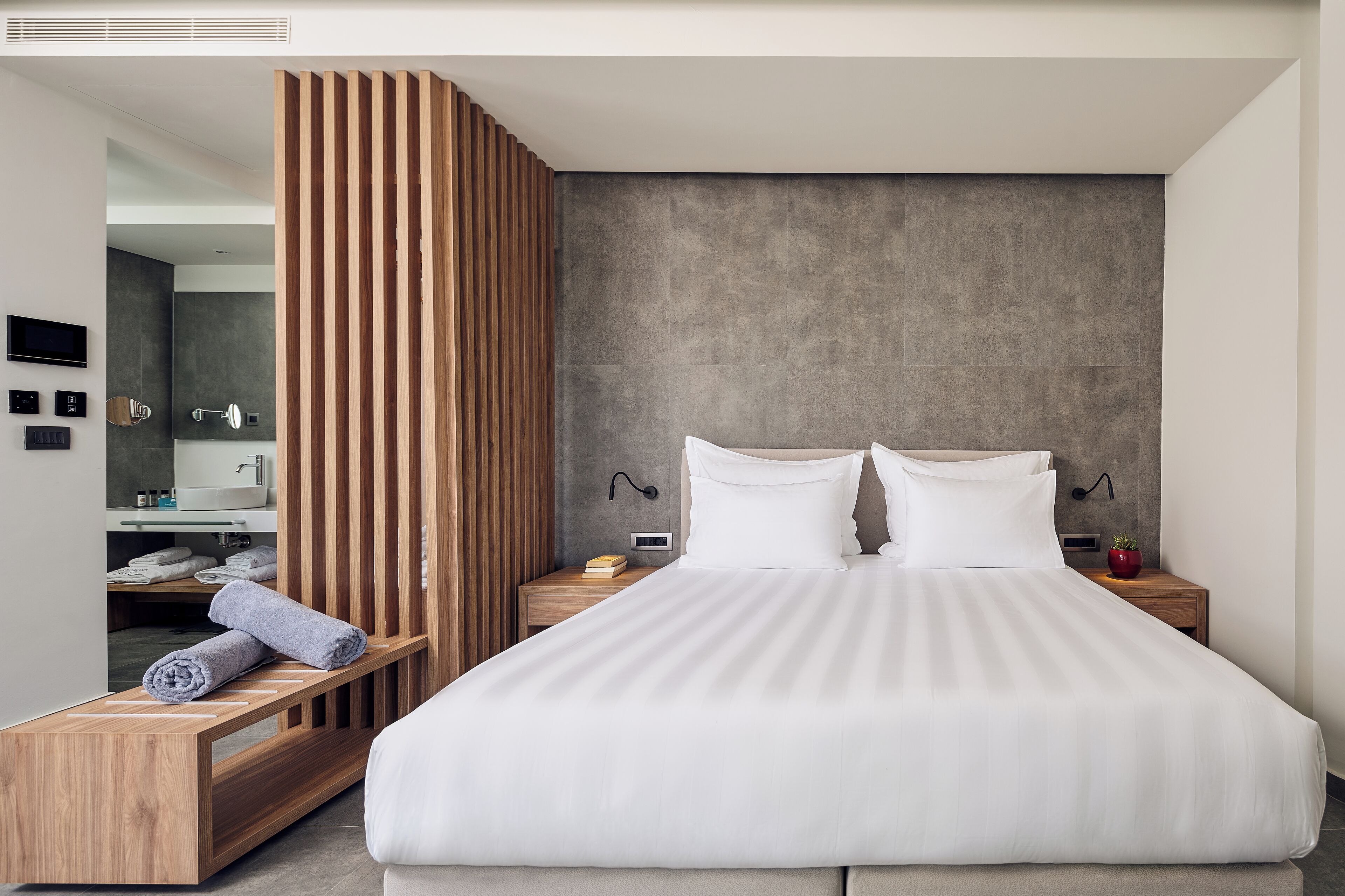 Ein modernes Schlafzimmer mit einem großen Bett, hölzernen Nachttischen, einer eingebauten Sitzbank und einer Wand mit strukturierter Grauoberfläche.