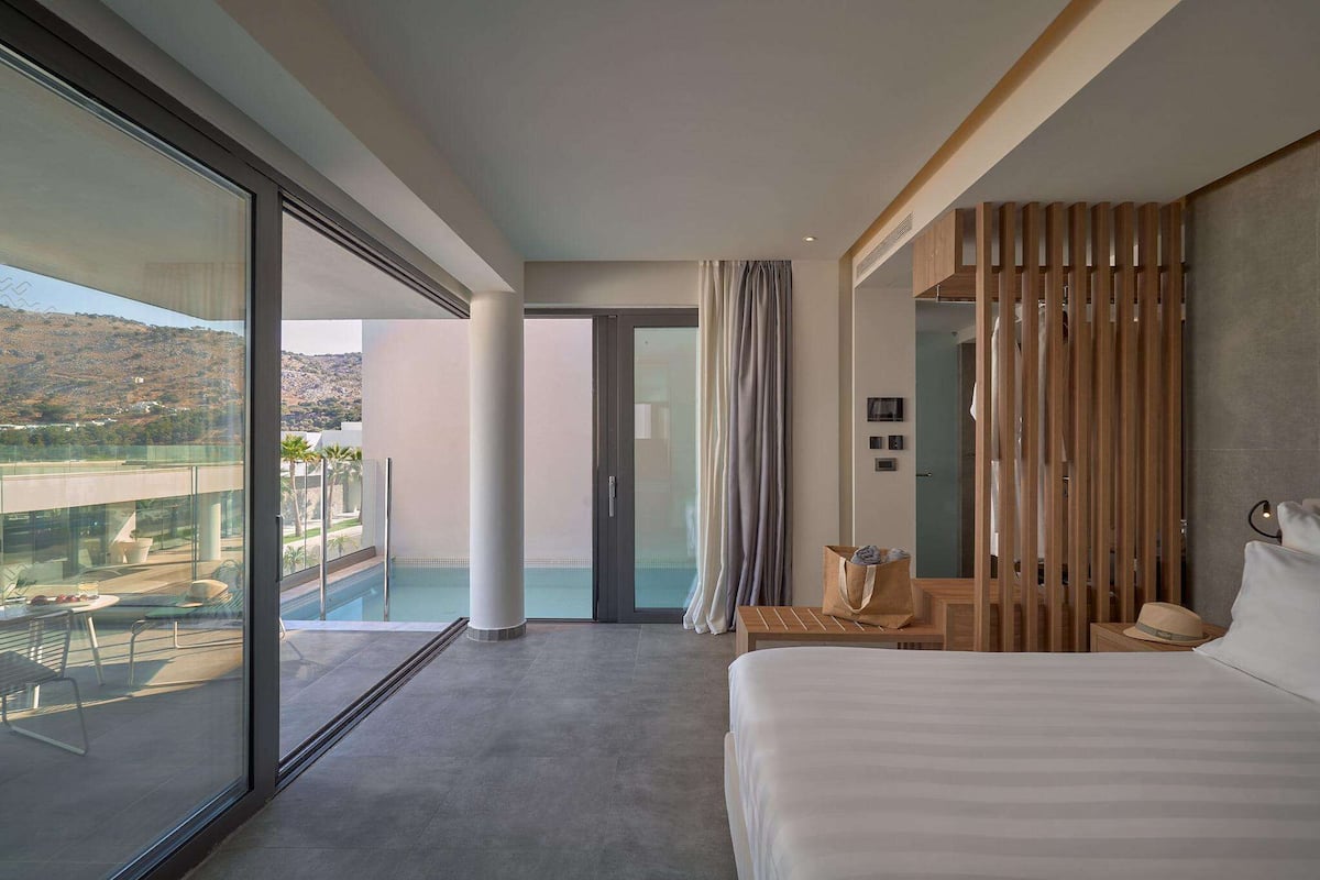 A modern hotel room with a large bed, a balcony with a pool view, and a sliding door leading to a shower area.