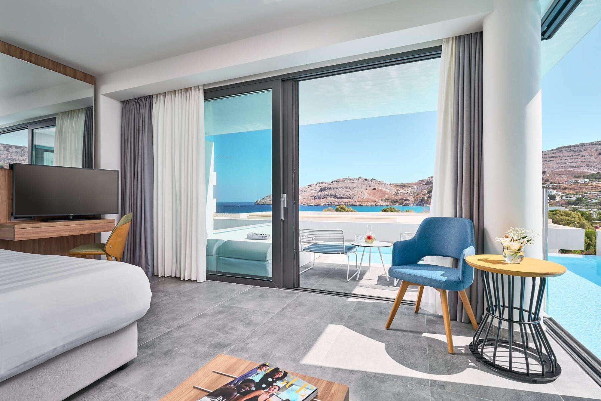 A modern hotel room with a large bed, a flat-screen TV, a balcony with a view of the sea and hills, and a small table with a vase of flowers.