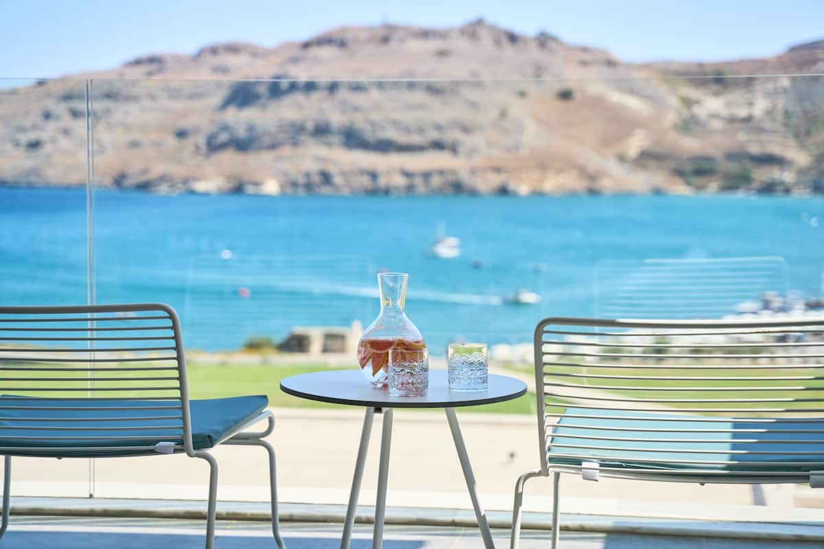 A balcony with two metal chairs and a small round table, overlooking a coastal area with clear blue water and distant hills.