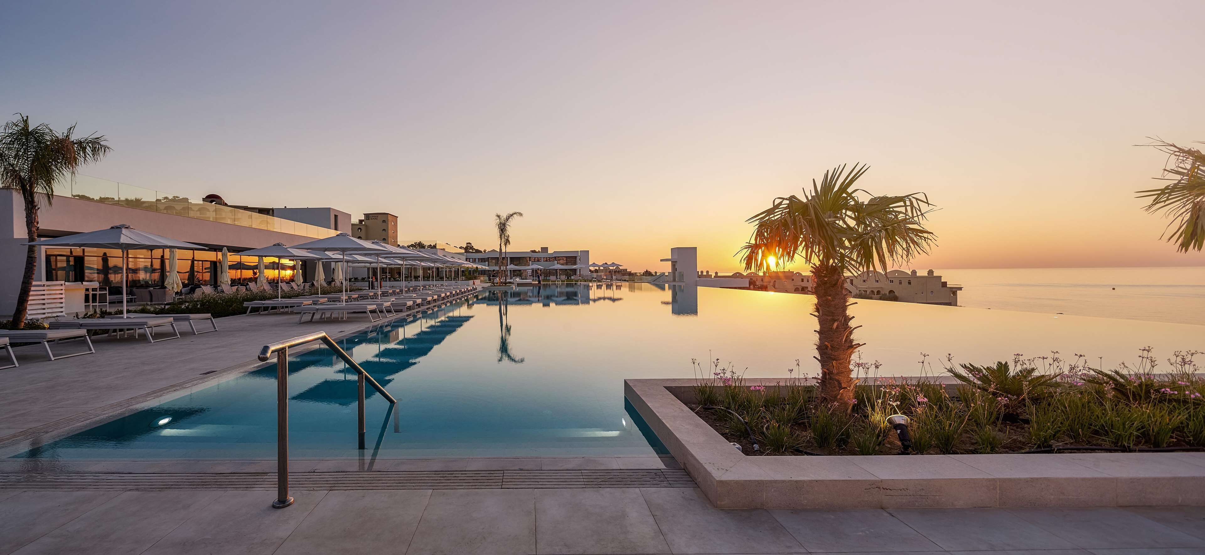 A pool area with lounge chairs and umbrellas, a clear view of the sunset, and a calm water body.
