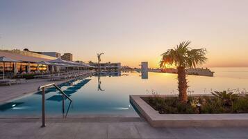 A pool area with lounge chairs and umbrellas, a clear view of the sunset, and a calm water body.