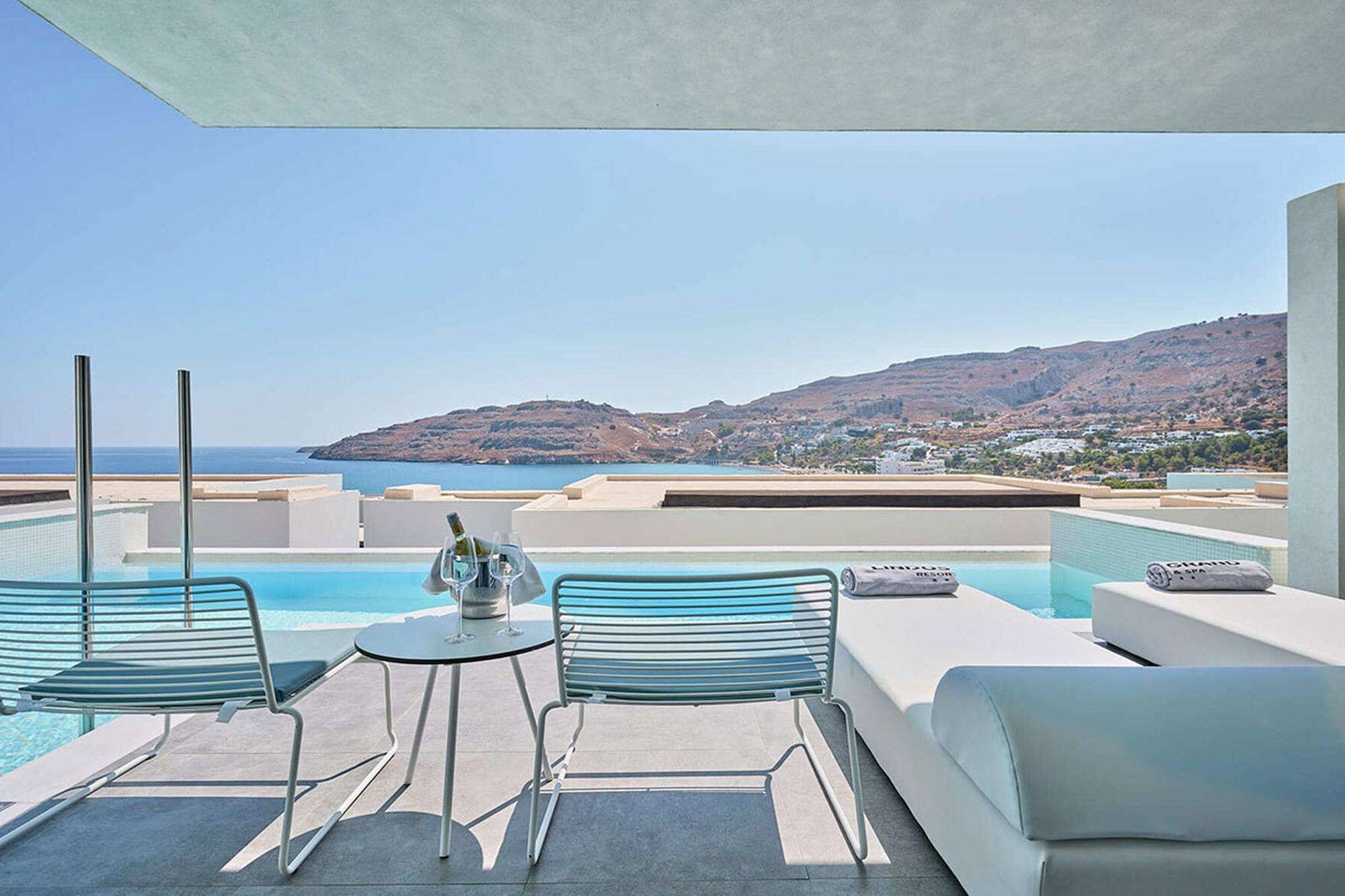 A rooftop pool area with a view of the sea and hills, featuring a table with chairs and a bottle of wine.