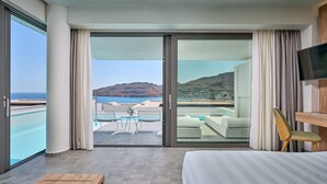 Habitación de hotel con una cama grande, una mesita de noche de madera, un televisor y un balcón con piscina y vistas al mar y a las colinas.