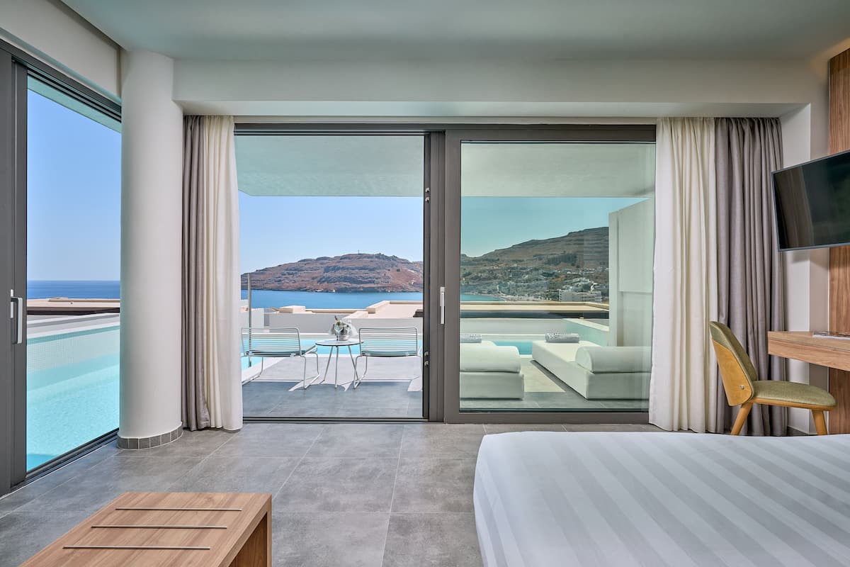 A hotel room with a large bed, a wooden side table, a TV, and a balcony with a pool and a view of the sea and hills.