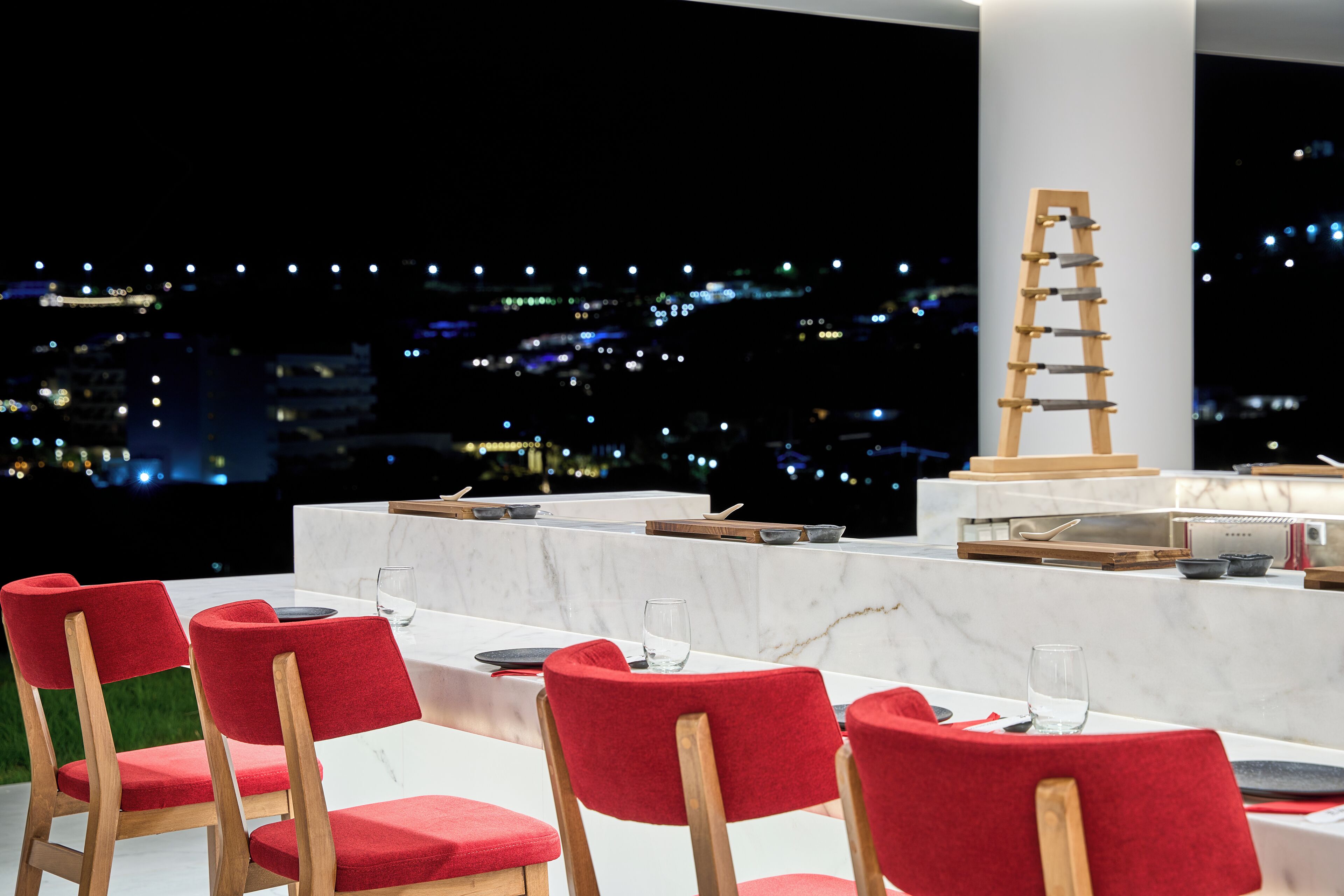 Uma sala de jantar com cadeiras vermelhas estofadas, uma mesa de mármore e vista para o horizonte noturno.