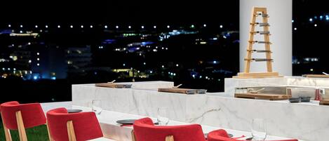 A dining area with red upholstered chairs, a marble table, and a view of a cityscape at night.