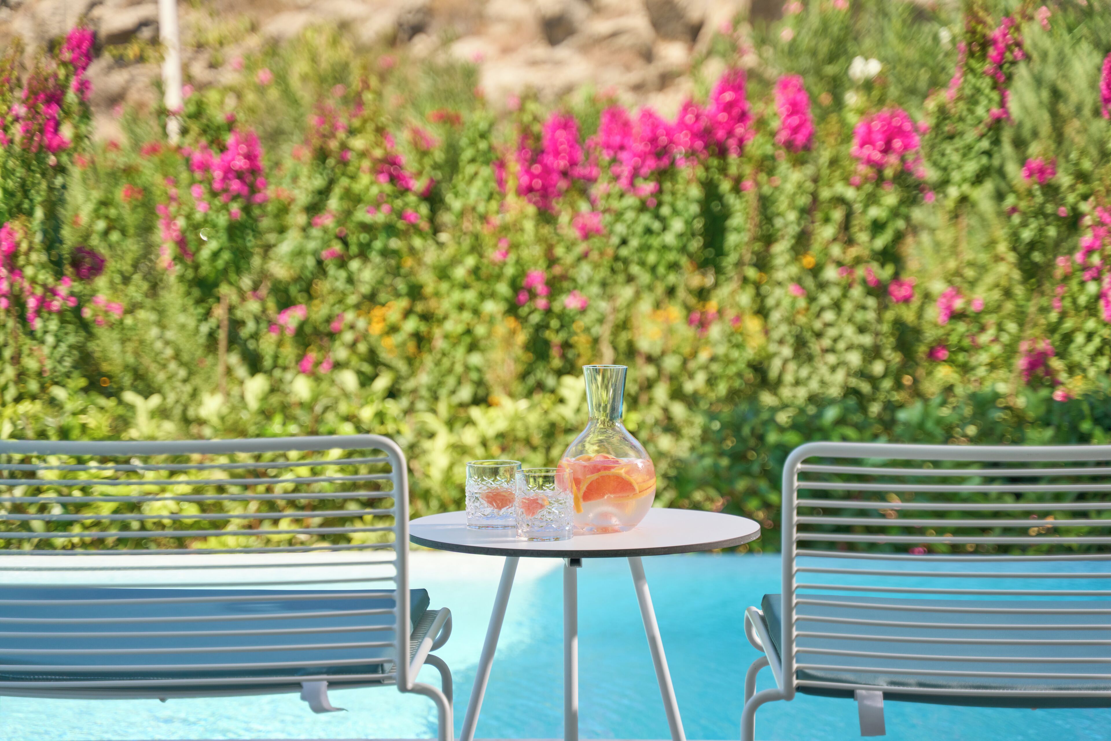 Un espace aménagé au bord de la piscine, avec deux chaises en métal et une table, une carafe en verre et deux verres.
