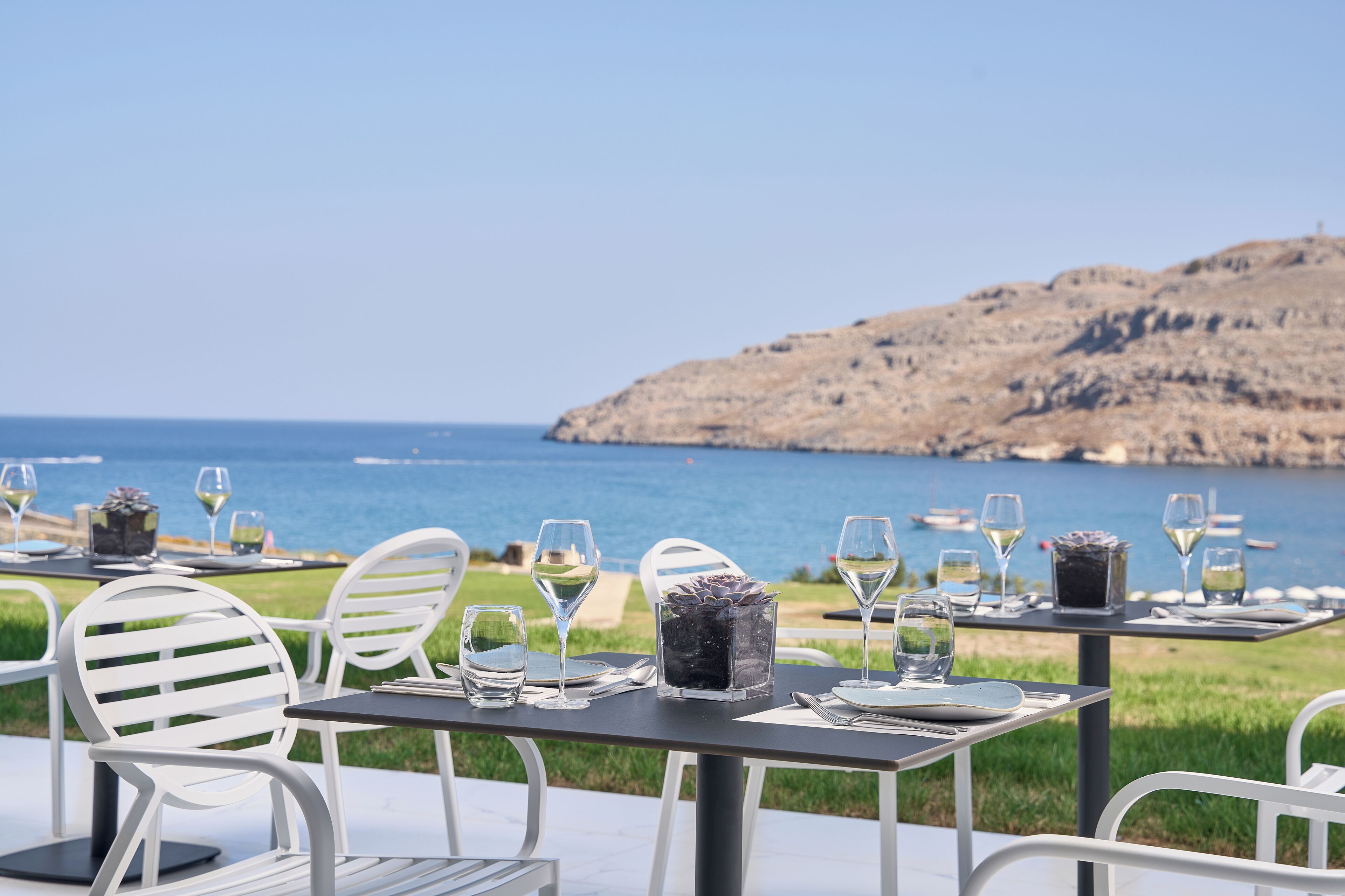 A table set for dining with chairs, a view of the sea, and a rocky hillside in the background.