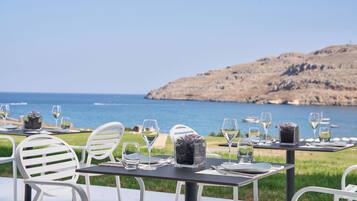 A table set for dining with chairs, a view of the sea, and a rocky hillside in the background.