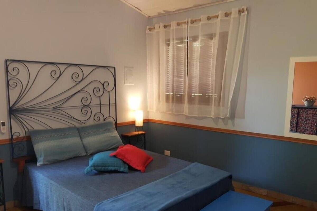 Ein Schlafzimmer mit Bett, Kissen, einem Nachttisch mit Lampe, einem Fenster mit durchsichtigen Vorhängen und einem kleinen Regal mit einer Pflanze.