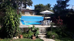 Une piscine aux parois bleues, entourée d’un jardin où poussent différentes espèces de plantes, et agrémentée d’un parasol blanc.