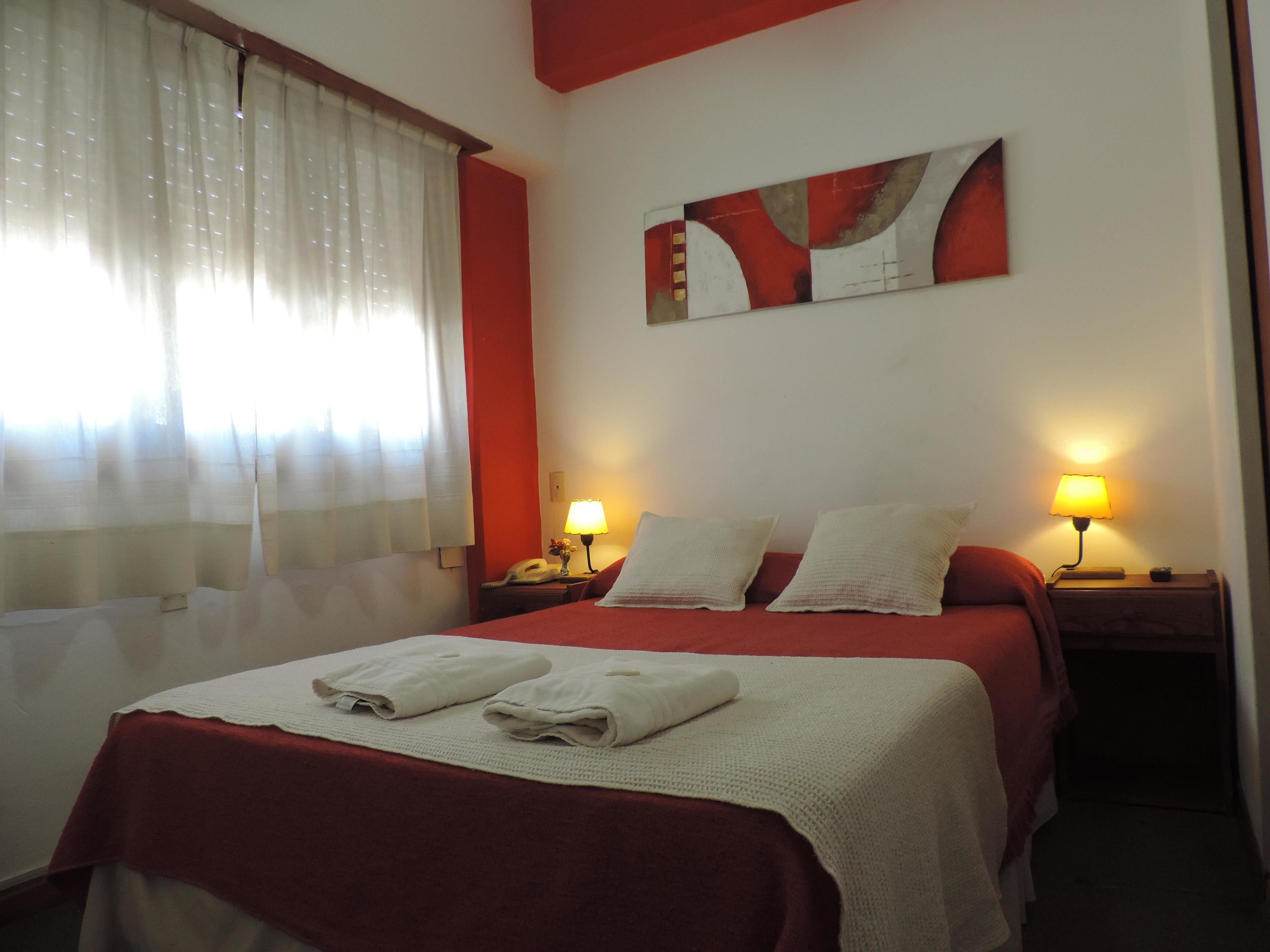 A hotel room with a bed, two bedside lamps, a painting on the wall, and a window with curtains.