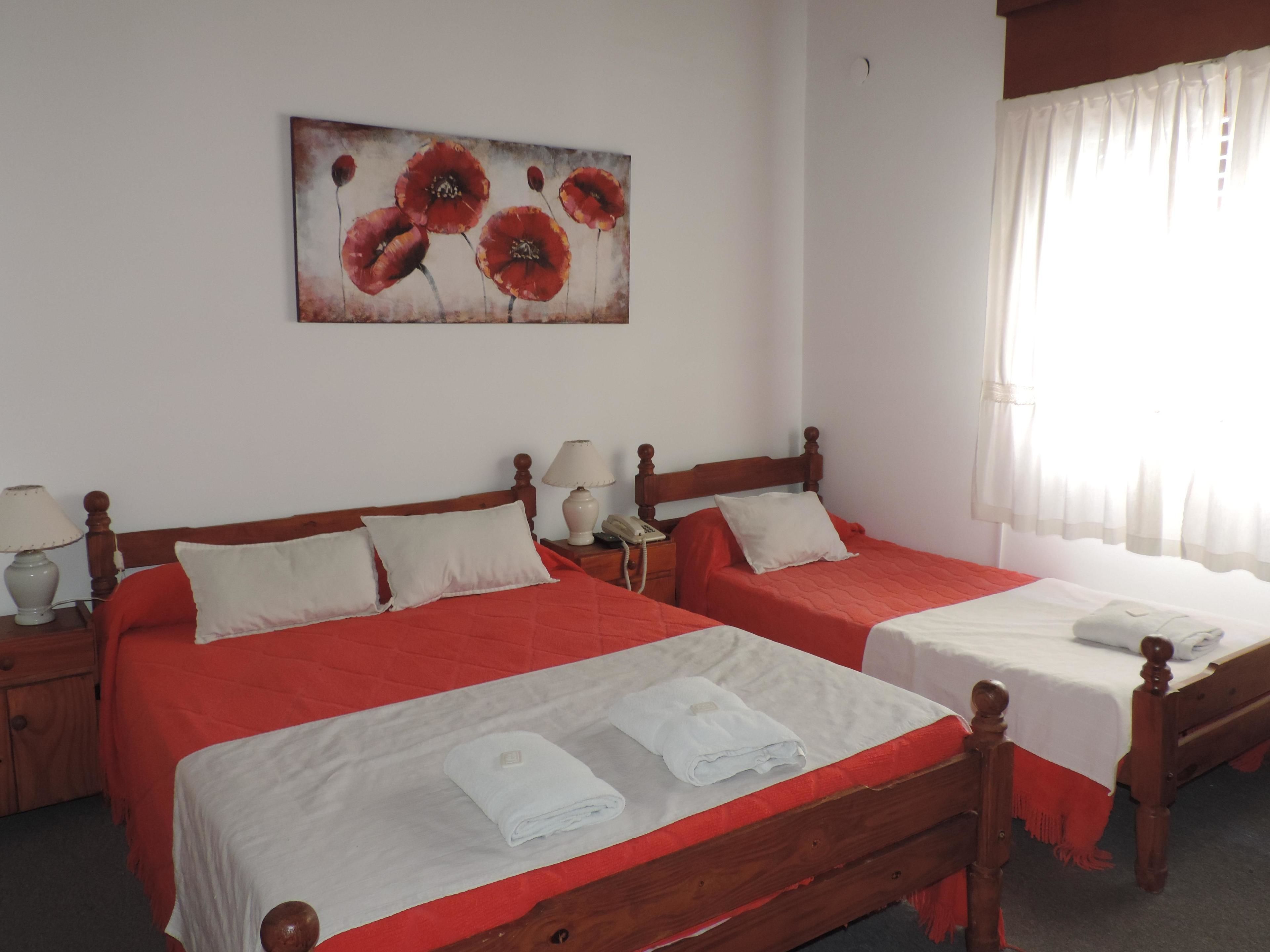 Une chambre d’hôtel avec deux lits, chacun doté d’une tête de lit en bois, de draps blancs et d’éléments décoratifs rouges. Un tableau représentant des fleurs rouges orne le mur, ainsi qu’une lampe de chevet et une fenêtre avec des rideaux légers.