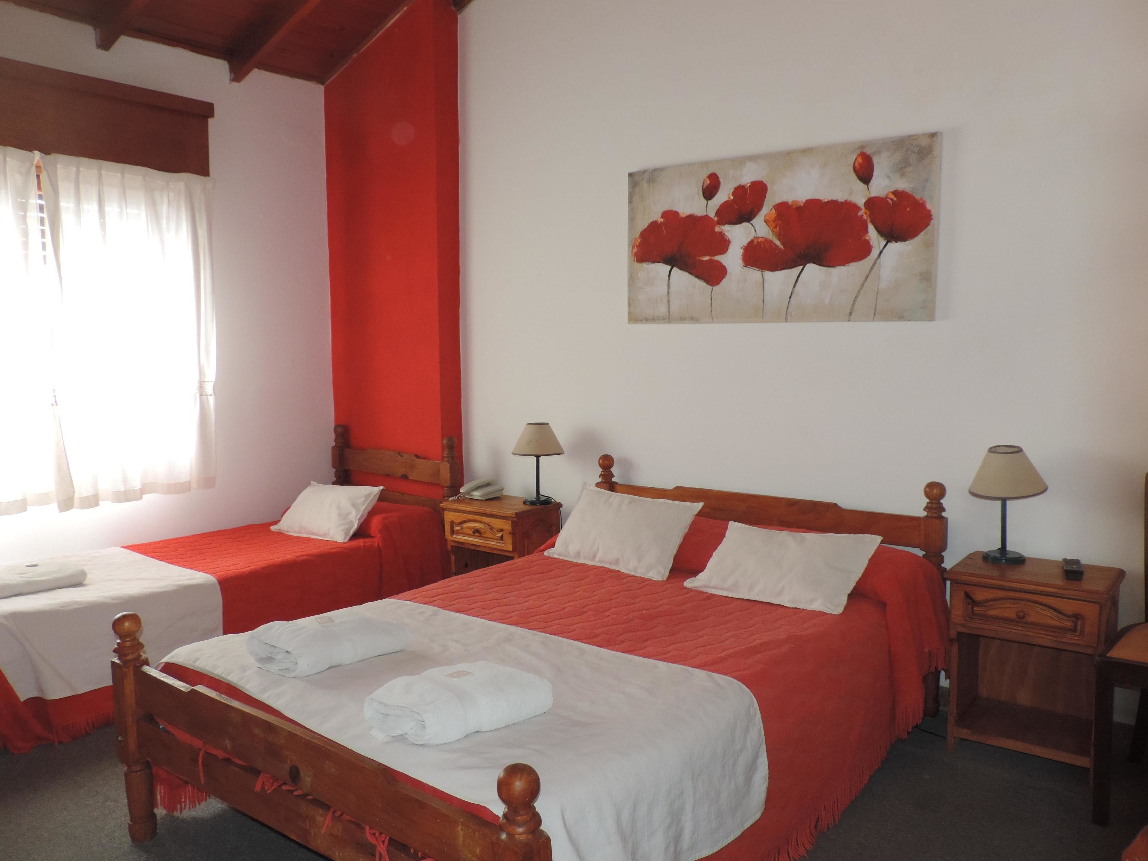 A hotel room with two single beds, a wooden headboard, a painting of red flowers, and a window with sheer curtains.