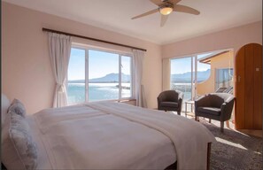A hotel room with a large bed, two chairs, a ceiling fan, and a view of the sea.