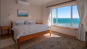 A bedroom with a large bed, a view of the sea, a ceiling fan, and a painting on the wall. - Bikini Beach Beauty (Cape Town)