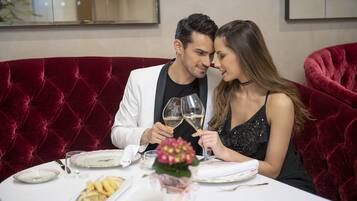 A couple dining at a table with a red velvet couch, white tablecloth, and a small vase of flowers.